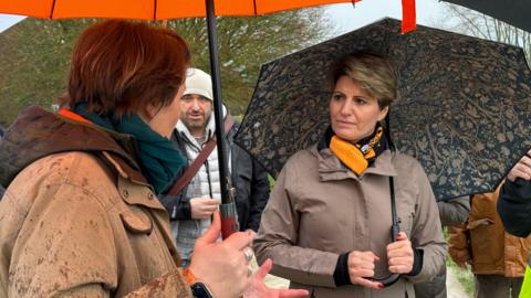 Two women are talking - the one on the left has her back to the camera - while the other - Emma Hardy, who has short brown hair, is looking at her. They are both holding umbrellas and wearing waterproof, brown coats.