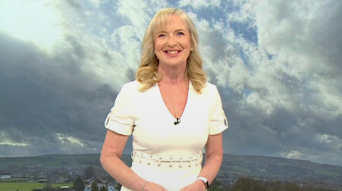 Kirkwood smiling at the camera in a cream dress holding her hands together in front of her stomach. There's a green screen background behind her showing rolling hills with a cloudy sky.