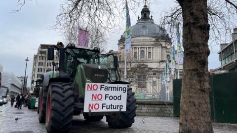 A green tractor is parked on a Westminster pavement under a grey winter sky next to a bare tree and in front of the QEII Centre. On the front of the tractor is a protest placard that reads No Farming, No Food, No Future. Pedestrians pass by.