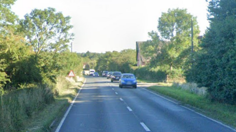 A Google Street View screenshot of a two-lane stretch of the A140 road in the UK, showing oncoming traffic led by a blue hatchback, with lush green hedges, mature trees, and a triangular road sign lining the countryside roadside under a bright sky.