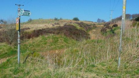 A patch of grassy, peaty, overgrown moorland, with a path winding upwards towards two hills visible, along with signs directing walkers