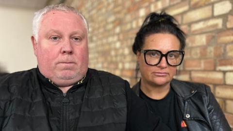 A man with grey hair wearing a black jacket sits next to a woman with dark hair, dark framed glasses and wearing a black t-shirt and jacket