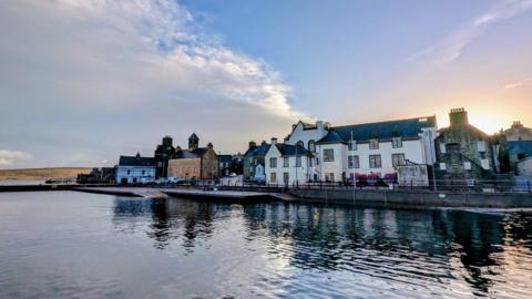 Buildings on the water's edge in Shetland with the Sun rising  behind and blue cloudy skies.