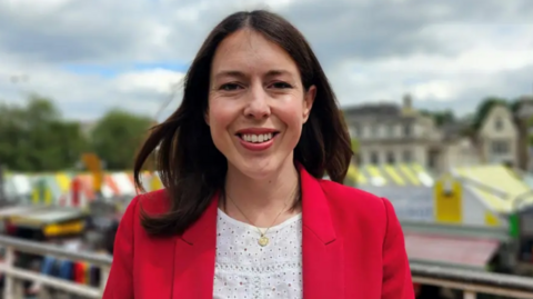 Alice MacDonald smiles directly at the camera as she is photographed outside, with Norwich market visible in the background. She is wearing a red jacket and white blouse, with gold necklace. She has shoulder-length brown hair.  