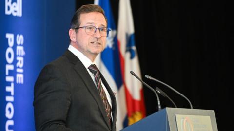 Yves-François Blanchet, in glasses and a suit, stands at podium