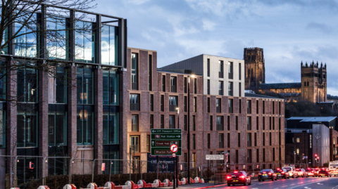 Nearly finished buildings on the Milburngate site. The multi-storey blocks are mostly made of glass and brown bricks. Durham Cathedral is shining in the distance as cars travel down Framwellgate towards the city centre. The site is fenced off. 
