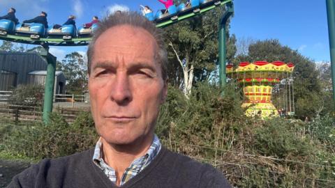 Close up of Rick Turner. . He is looking into the camera with a frown.He is standing outdoors in front of a rollercoaster. People are in carts on the rollercoaster. Some people have their arms in the air. A yellow swing ride is in the background behind a hedge.