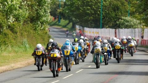 A group of motorcyclists on classic racing bikes with yellow plates in the front on a section of the Billow Circuit, which has barriers to the right and a grass bank to the left.