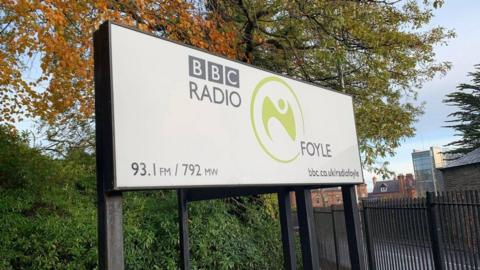 A sign outside of ˿ Radio Foyle. It has the wave lengths and website on the sign. It has been taken on a sunny autumnal day