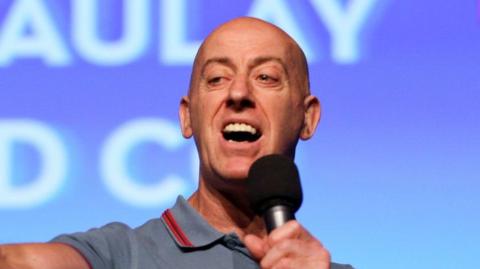 Gary Little speaks into a microphone - he is bald and wears a light blue polo shirt.