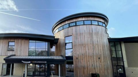 The front of a wooden-clad building including a tower shaped two-storey section and glass fronted entrance with The Roundhouse in white lettering above it.