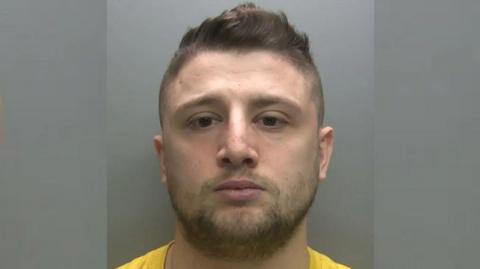 Mugshot of Jacob Corrie who has a round face and brown eyes. He has a short back and sides haircut and a small amount of facial hair. He is wearing a yellow T-shirt. 