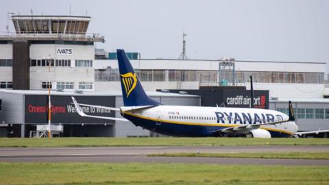A Ryanair plane, with white body, blue text and a blue and gold harp logo on its tail fin, taxis in front of the terminal building at Cardiff Airport. Grass and tarmac can be seen in the foreground.