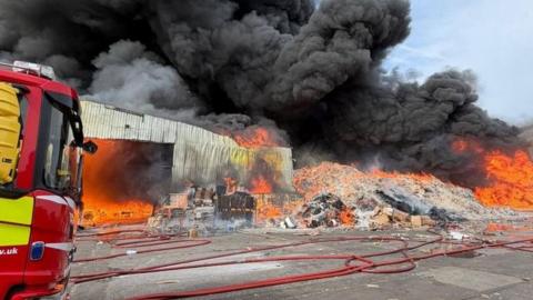 The front of a fire engine can be seen on the left of the image, with a large pile of cardboard on fire and large plumes of smoke billowing into the sky. A single-storey building can also be seen ablaze.