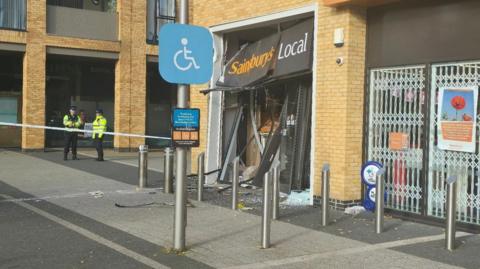 The outside of a Sainsbury's Local store in Brooklands Square. The front of the shop has been damaged with the doors warped and smashed glass across the floor in front of the shop. There is a police cordon in place and two police officers are standing inside the cordon.