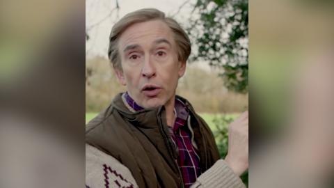 A man, Alan Partridge, wears a checked shirt and a jacket with patterned sleeves. He is standing outside.