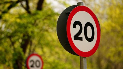 A picture of two signs both saying 20 on it. There is red and white signs. There is large trees and leaves in the background.
