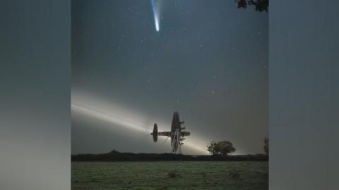 The bright blue, Comet Lemmon going across the night sky leaving a light train behind it above the Lancaster bomber sculpture, with a gray light in the sky behind it