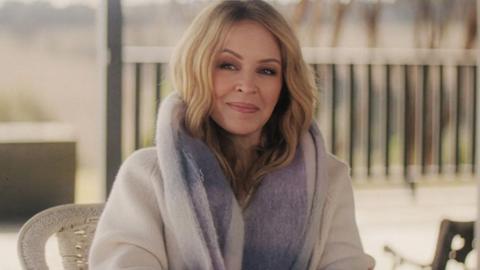 Kylie Minogue sitting down in a white coat and purple scarf