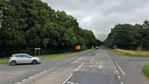 A silver car on Swaffham Heath Road waits to cross the A1303 at the crossroads.