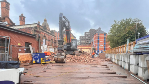 A fenced off building site where a large digger is parked on a pile of bricks and rubble.  Leicester's Victorian rail station can be seen in the back ground.