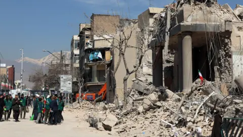 Rubble from a damaged building is visible beside a road with a group of people standing beside it