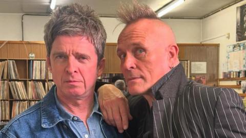 Noel Gallagher is standing next to John Robb in a vinyl shop. John is leaning on Noel's shoulder and both men are staring directly at the camera. Liam is wearing a denim jacket and denim shirt while John is wearing a pinstripe suit and shirt with the collar up. 