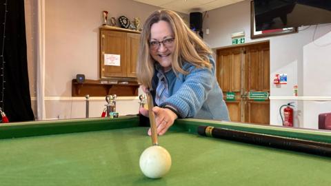 Jennifer Barton with long blonde hair and glasses, wearing a blue cardigan and holding a snooker cue whilst leaning on the table. She is about to hit the white ball which is near the centre of the snooker table. There is another cue resting on the table. In the background, there are wooden doors and white walls. Some trophies sit atop a wooden cupboard.