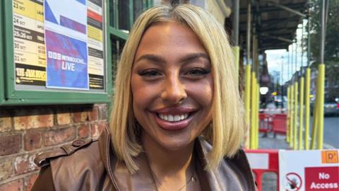 Maddie Juggins outside the BBC Radio 1Xtra Introducing gig smiling at the camera. She is wearing a brown leather jacket, and behind her are yellow construction poles with a red "no access" sign and barricade.