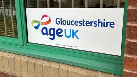 A sign in a window says Gloucestershire Age UK. The window frame is green.