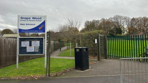 A school sign with a white background and blue writing. It is behind a green gate.