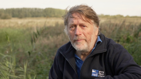 A man wearing a dark fleece over a blue polo shirt. He is looking into the camera. There is grassland in the background.