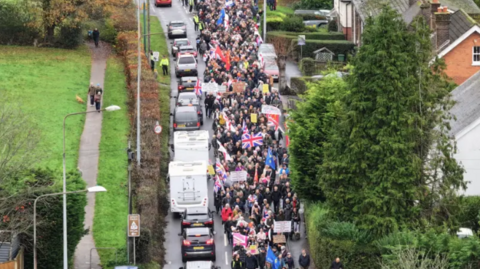 A drone shot of a protest held in Crowborough, East Sussex on 16 November over plans to house asylum seekers at an army site in the town. Hundreds of people can be seen walking on the streets with flags and banners. Cars are driving alongside the walkers. 