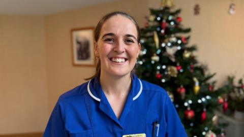 A woman wearing a bright blue nurse uniform looking towards the camera and smiling. She has brunette hair and is standing in front of a Christmas tree.
