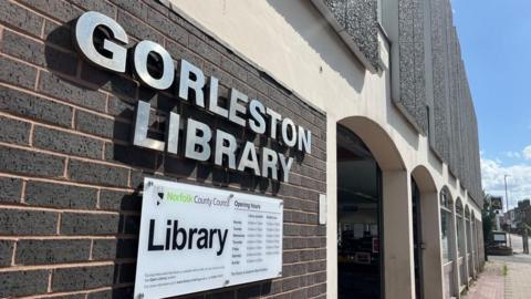Chrome signage stating "Gorleston Library" and the library opening hours on a tablet below, bearing the logo and name of Norfolk County Council. The building is a 1970s design, featuring brown bricks, concrete pillars and arches, several infilled with aluminium-framed windows, and flint-faced concrete panels on the upper storey.