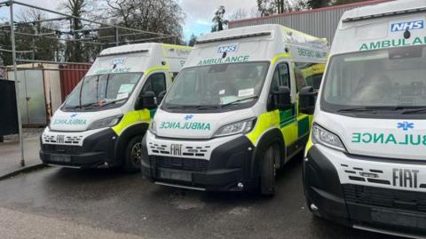 Three new FIAT white ambulances are parked up at an industrial facility. The white ambulances have green and fluorescent yellow markings with the NHS blue logo on the head of the vehicles.