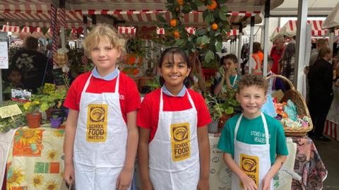 Three children stand in a row wearing white aprons in front of market stalls with fruit and plants on them.