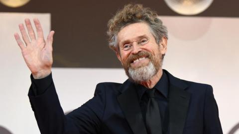 Willem Dafoe wearing an all black suit waving with one hand to the cameras on the red carpet.