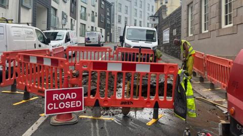 A Jersey Water engineer wearing yellow high-vis clothing works at the site of a burst water main on Pier Road in St Helier. Safety fencing has encircled the site and a road close sign is visible.