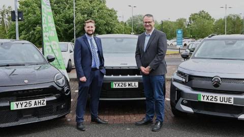 Two men stand in front of a black electric car, with other models to either side, in a car park. The man on the left has brown hair and a beard and is wearing a blue suit and blue and white striped tie. The man on the right has short grey hair and wears glasses, a grey blazer and blue jeans.