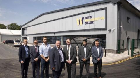 A group of seven men in suits stand in front of a metal clad building which has a sign saying City of Wolverhampton College