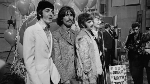 The Beatles post for a photo. Their clothing is from 1967. The photo is black and white. Some photographers are visible in the side of the image. 