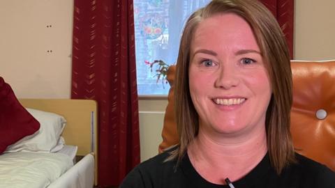 A woman smiles at the camera. She has shoulder-length brown hair and blue eyes, and is wearing a black top. Behind her is a bed and a window with red curtains