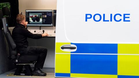 The image shows a police van with an open door that says "POLICE" in blue and has blue and neon yellow chequered boxes underneath. Inside the van, on the left-hand-side, a man can be seen sat down in an office chair looking at a computer screen. Two men wearing black can be seen on the screen.