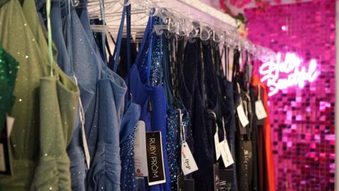 A colorful array of glamorous and sparkly prom dresses hung up in a shop they each have a tag on.