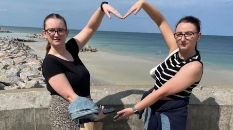 Megan and Lizzie stood by a beach and using their arms to create a heart