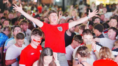 An England fan is standing up and sprawled her arms out wide among a sea of other England fans.