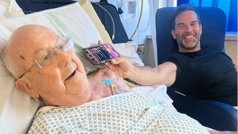 Gethin Jones on the right wearing a black t-shirt holding his phone up to his father's ear. The Spotify music app is visible on his phone suggesting he is playing him music. He is sat in a hospital room chair in front of a window with the blinds closed. His father, Goronwy, is in a hospital gown with glasses on listening to the music.