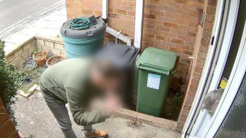 A man is stood at the door bent over talking to an elderly woman. He is wearing a green jumper. There are bins by the door and the woman is leaning out the door towards him to hear what he's saying.