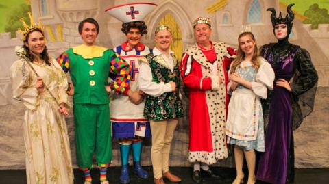 Seven people all dressed in pantomime costume standing on a stage and smiling. They are standing in front of a stage background featuring a castle scene.